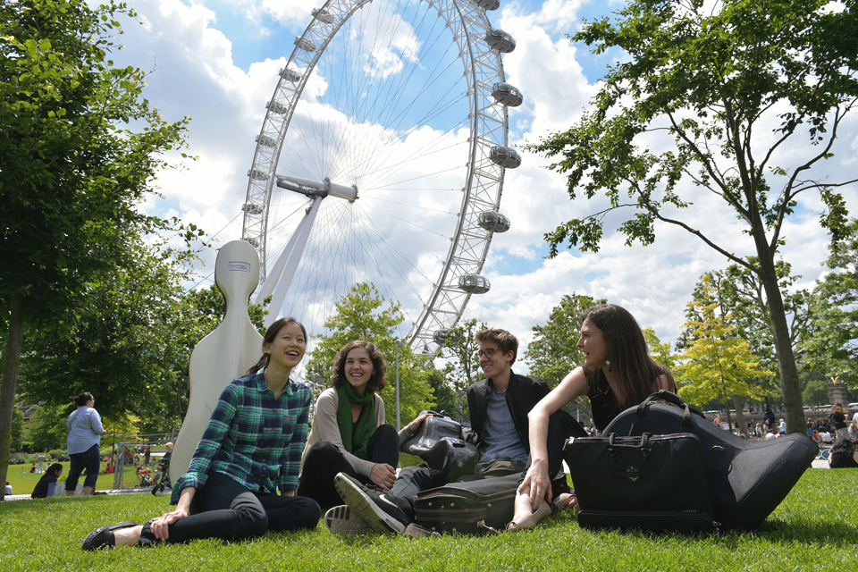 A group of students, smiling and chatting, sitting on the grass on Southbank, with the London Eye in the background.